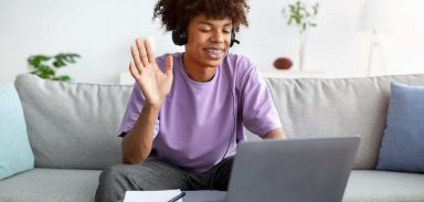 Happy black teenager in headphones greeting his friend or teacher online, waving at camera from home. Positive African American teen student having web conference, chatting to someone remotely