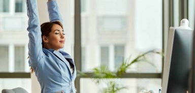 Young smiling businesswoman stretching with her eyes closed while working on a computer in the office.